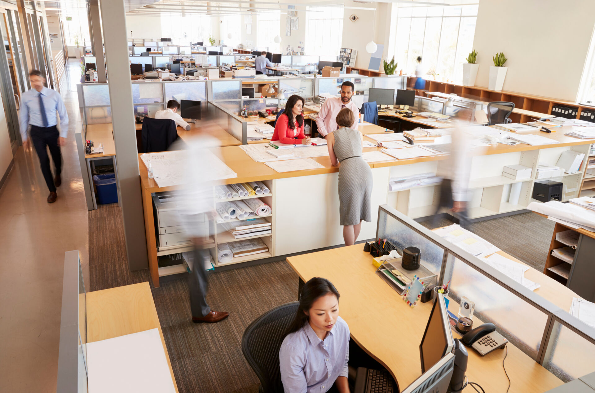 Employees moving through a busy open-plan office with desks, drawings, and shared workstations