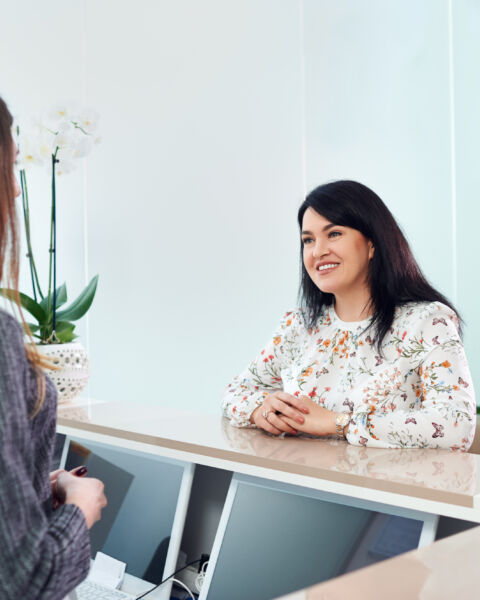 Mid adult woman with beautiful smile talking with young female receptionist, checking appointment in dentist clinic