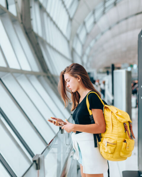Teen girl using smarthphone while waiting for international flight in airport departure terminal. Young passenger with backpack travelling on airplane. Teenager tourizm abroad alone concept.