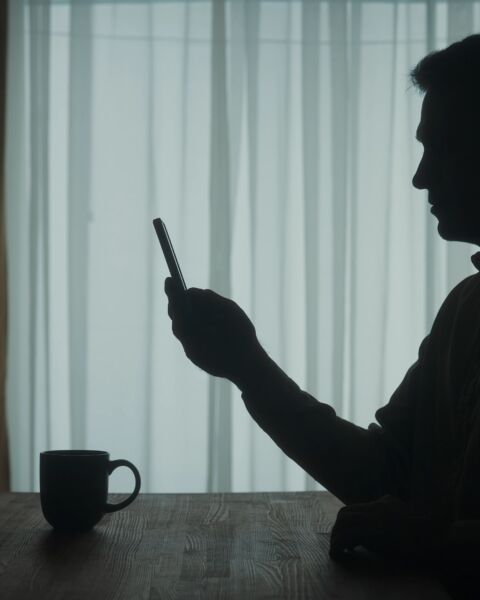 A man stands at a table with a phone in his hand, a cup of coffee stands nearby. A man flips through the tape, types a message, looks at photos, videos, talking on video call. The dark silhouette of a man in the living room at the table.